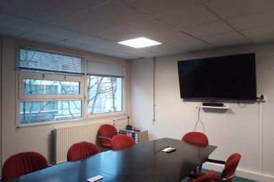 Salle de classe vide avec des rangées de tables et chaises, tableau blanc et écran au mur.