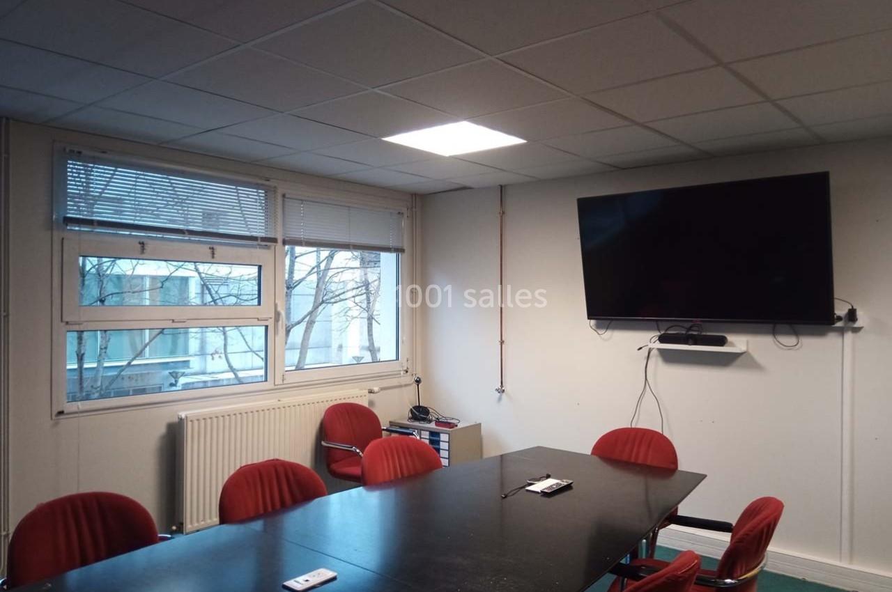 Salle de réunion avec table noire, chaises rouges, écran mural, fenêtre donnant sur des arbres et plafond éclairé.