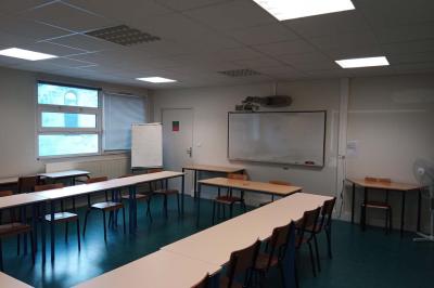 Salle de classe vide avec des rangées de tables et chaises, tableau blanc et écran au mur.