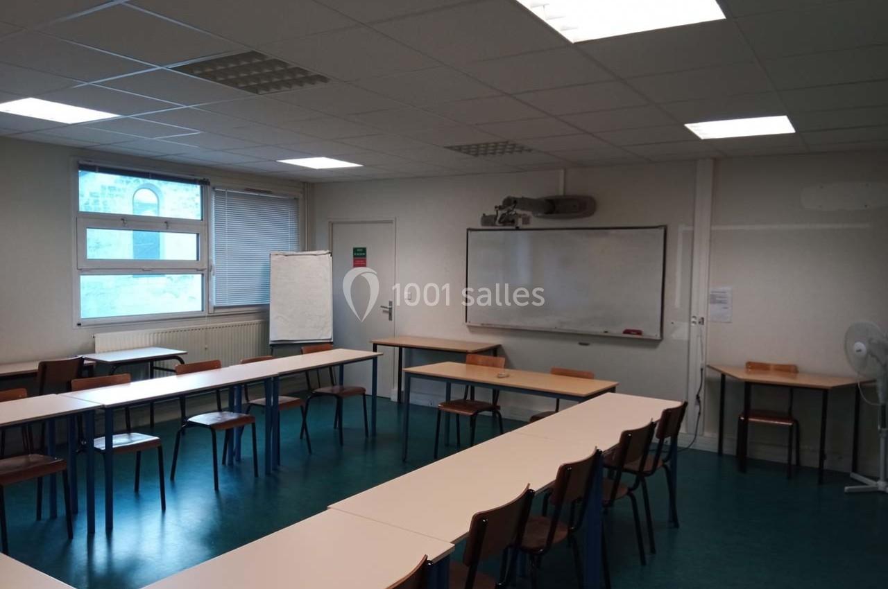 Salle de classe vide avec tables disposées en U, tableau blanc, vidéoprojecteur et fenêtres laissant entrer la lumière.