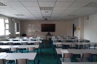 Salle de classe vide avec des rangées de tables et chaises, tableau blanc et écran au mur.
