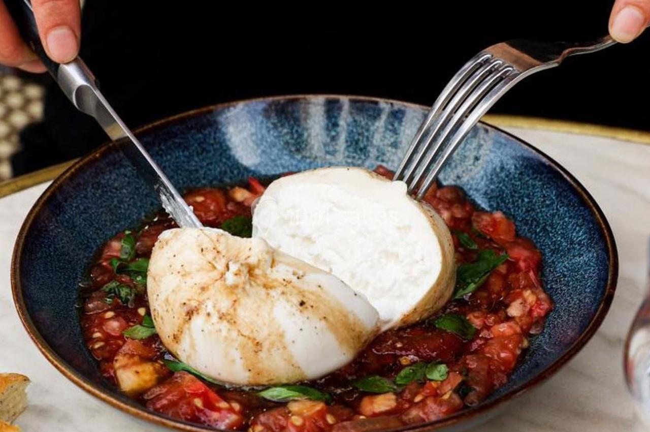 Une burrata coupée au couteau et à la fourchette, servie sur un lit de tomates concassées dans une assiette bleue.