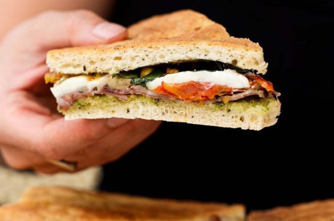 Sandwich focaccia garni de mozzarella, tomates, jambon et pesto, tenu dans une main.