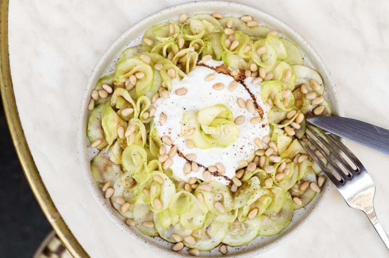 Assiette garnie de tranches de concombre, fromage frais, pignons de pin et épices, avec une fourchette et un couteau.