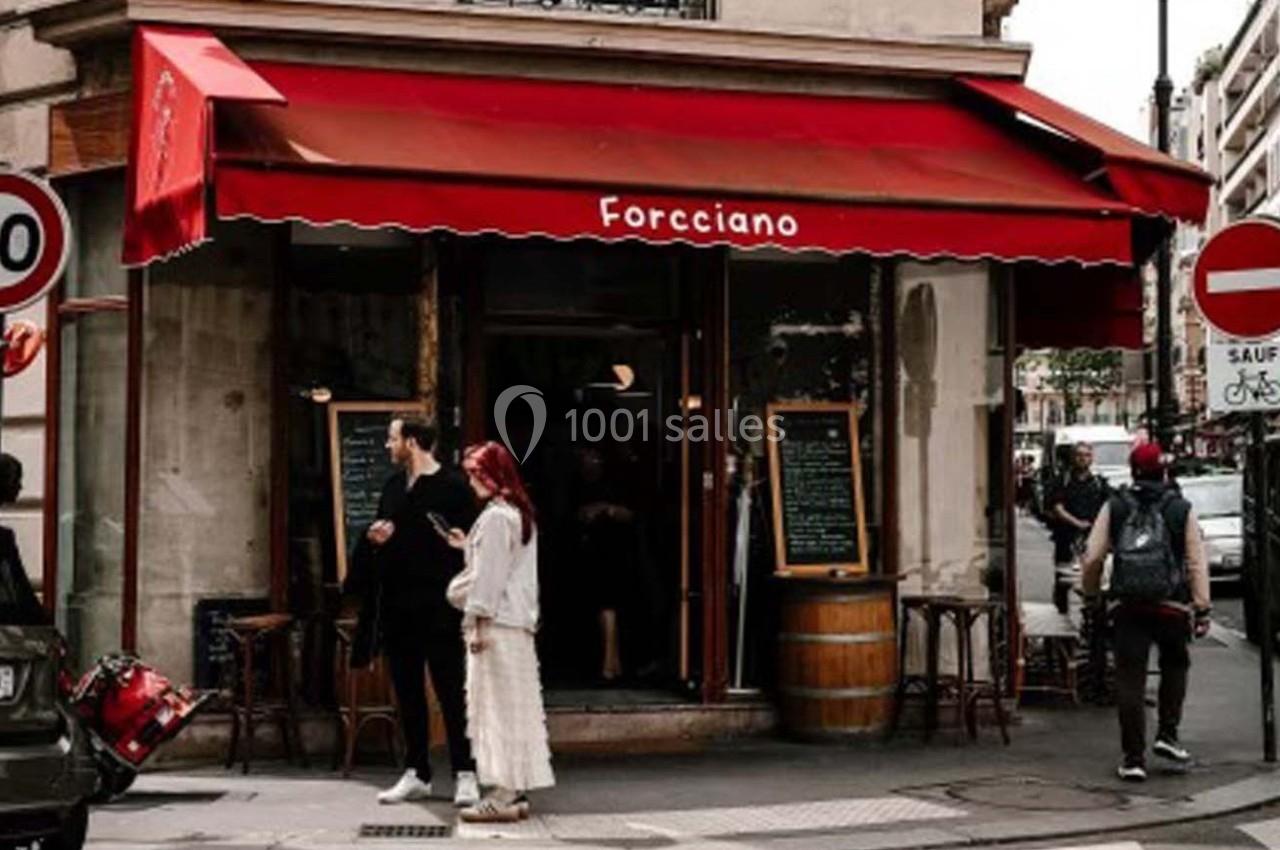 Façade d'un café avec une terrasse, une enseigne rouge et des passants devant, dans une rue urbaine.