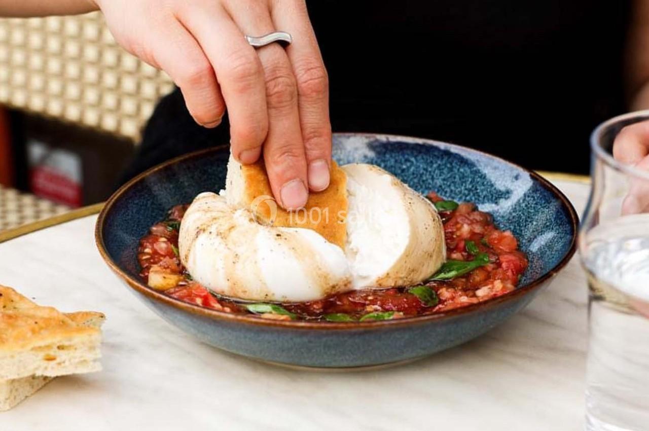 Une main trempe un morceau de pain dans une burrata servie sur un lit de légumes dans une assiette bleue.