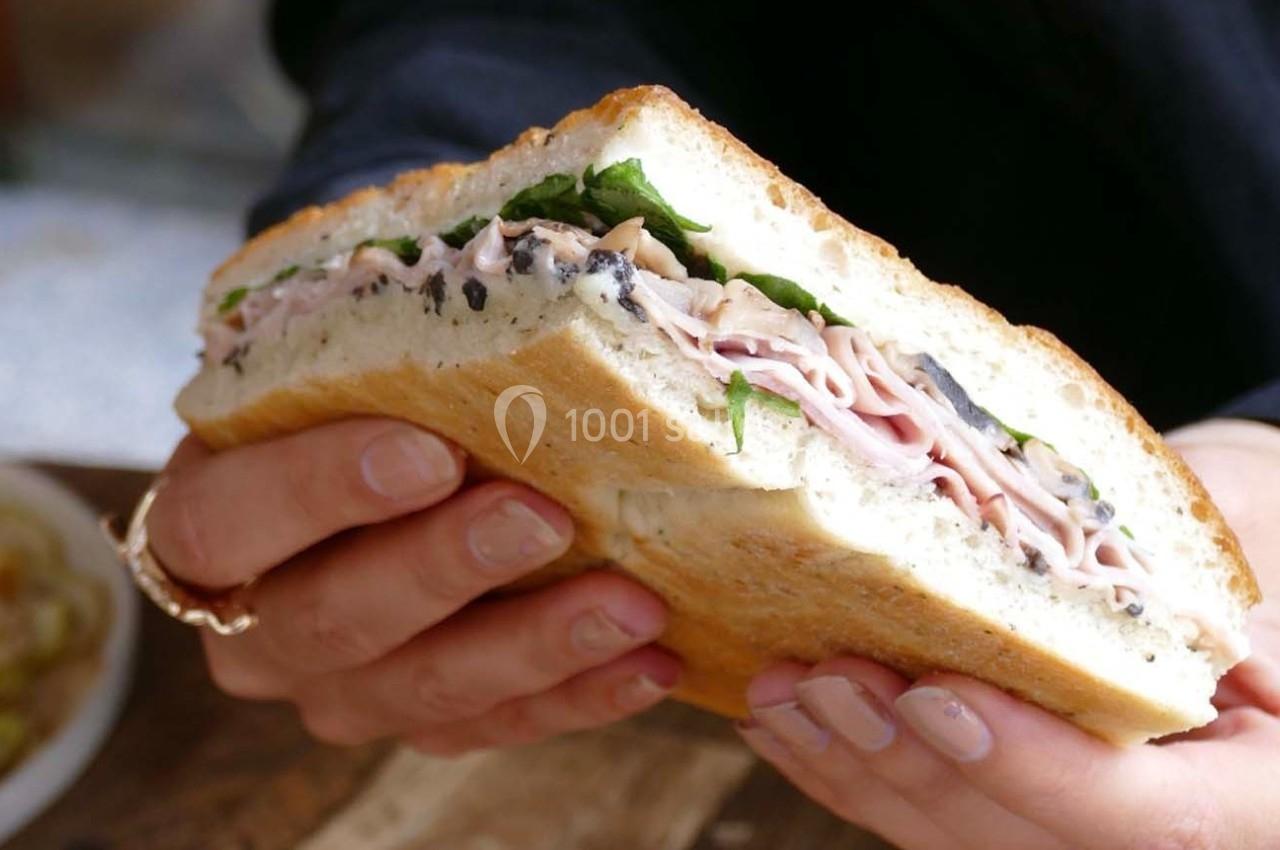 Un sandwich garni de jambon, salade et olives, tenu à deux mains sur un fond flou.