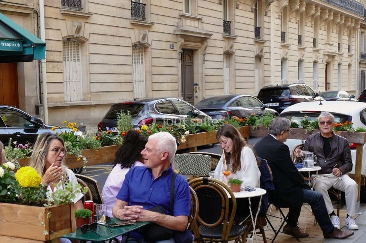 Des personnes discutent et prennent un verre en terrasse d’un café, bordée de jardinières fleuries, dans une rue urbaine.
