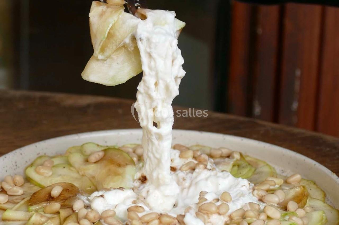 Tranche de pomme nappée de fromage crémeux, accompagnée de pignons de pin sur une assiette.