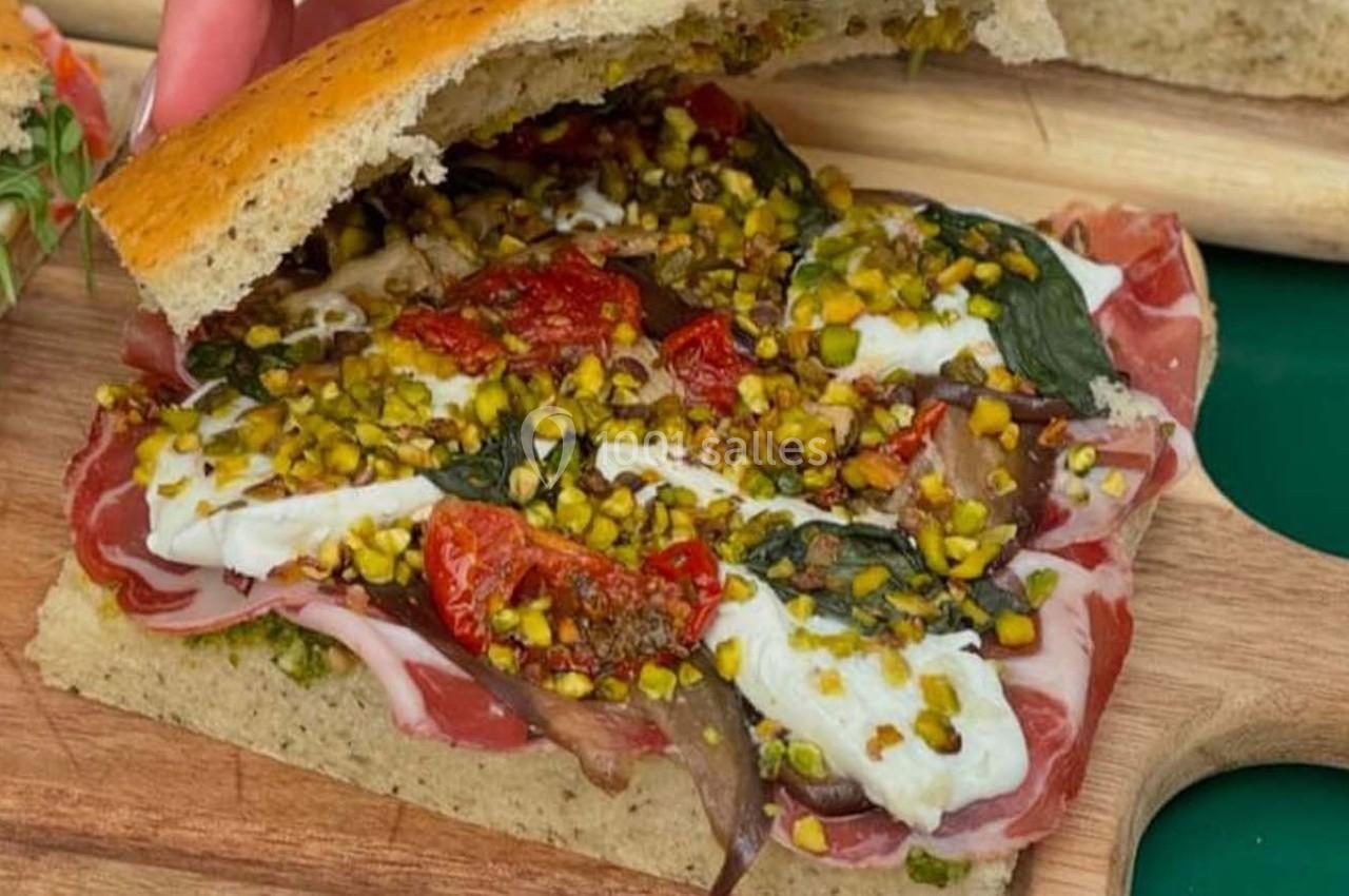 Sandwich ouvert garni de charcuterie, mozzarella, tomates séchées, pistaches concassées et feuilles de basilic.