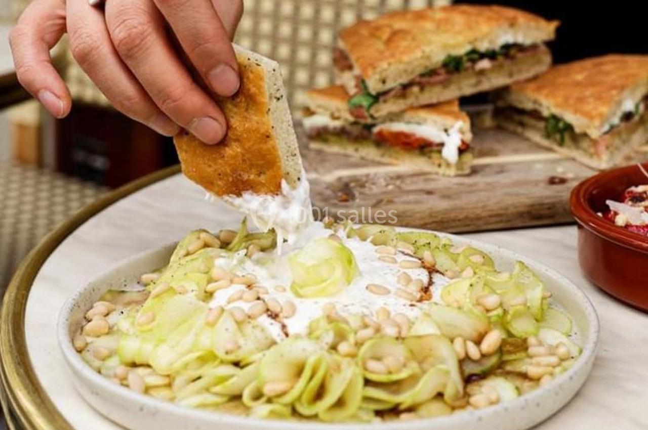 Une main trempe un morceau de pain dans une sauce blanche, à côté d'une assiette de salade et de focaccia.