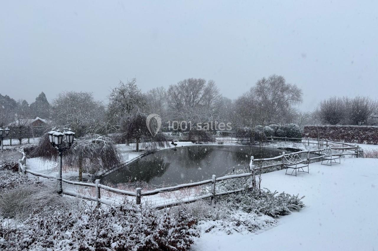 Étang entouré d'arbres et de buissons sous la neige, avec un lampadaire et des bancs dans un paysage hivernal.