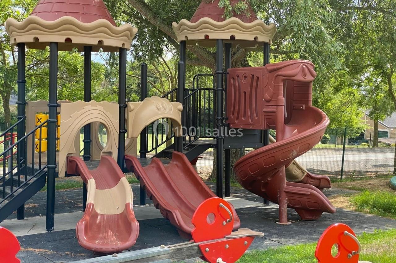 Structure de jeux pour enfants avec toboggans rouges et beige, située dans un parc arboré.