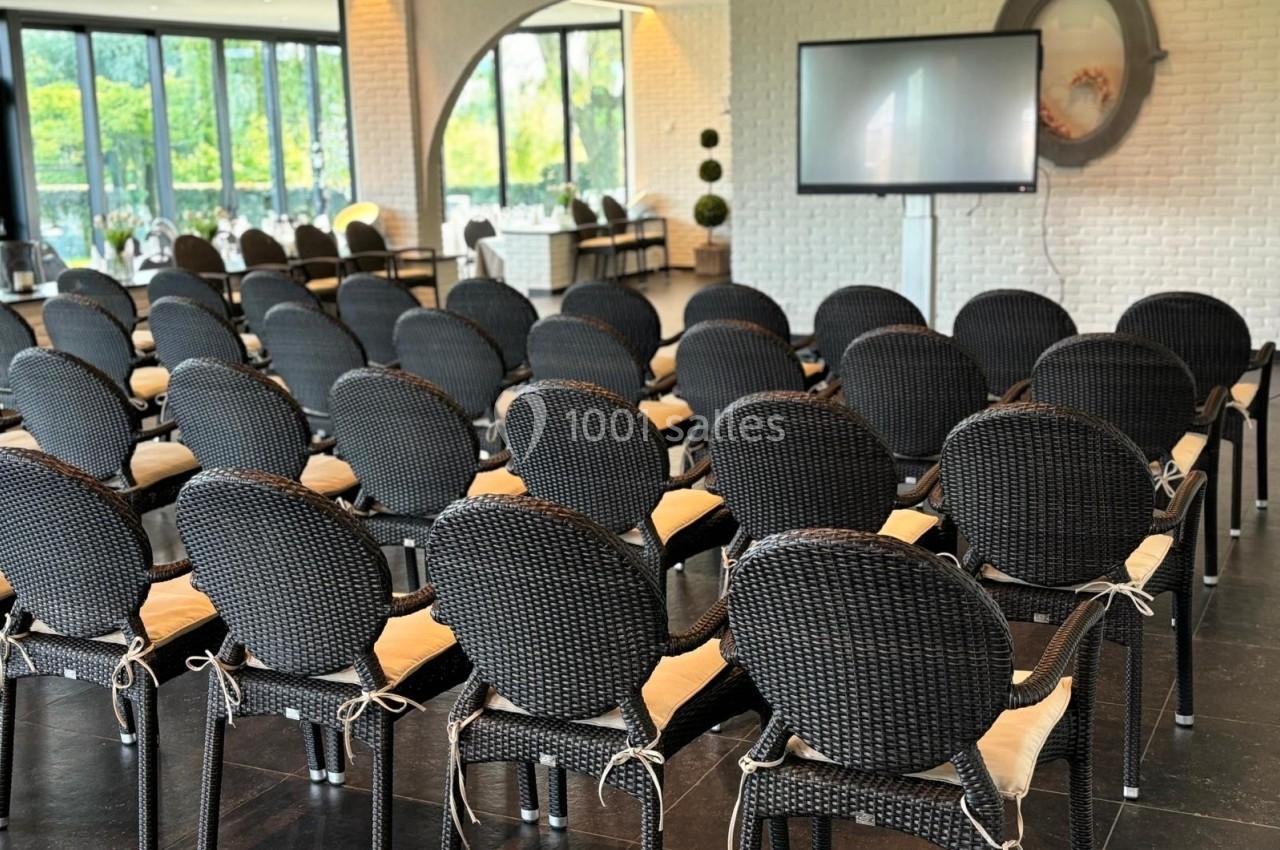Salle lumineuse avec des rangées de chaises noires en osier, un écran et de grandes fenêtres donnant sur un espace vert.