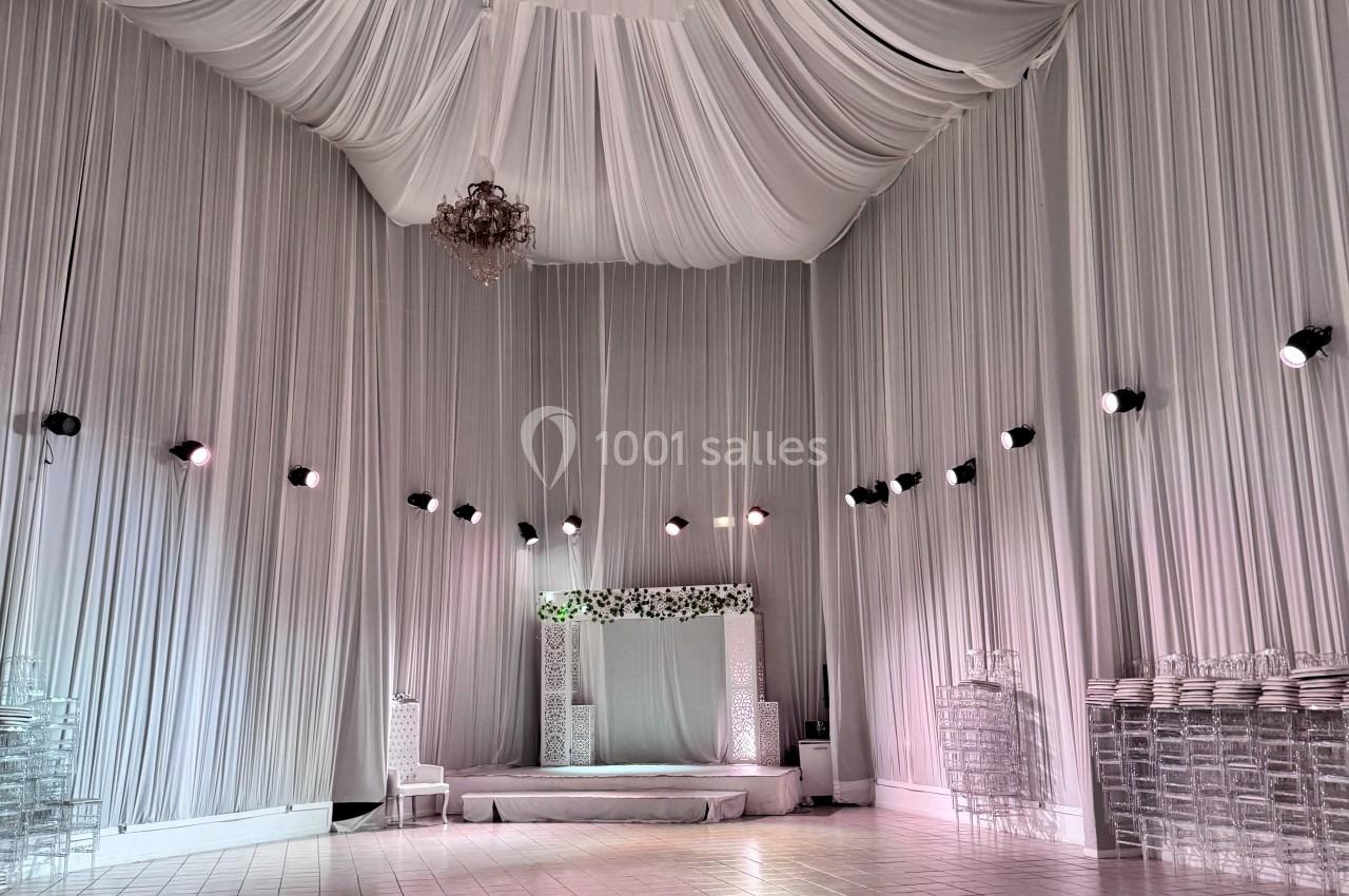 Salle de réception décorée de tentures blanches au plafond, avec une estrade ornée de fleurs et des chaises transparentes.