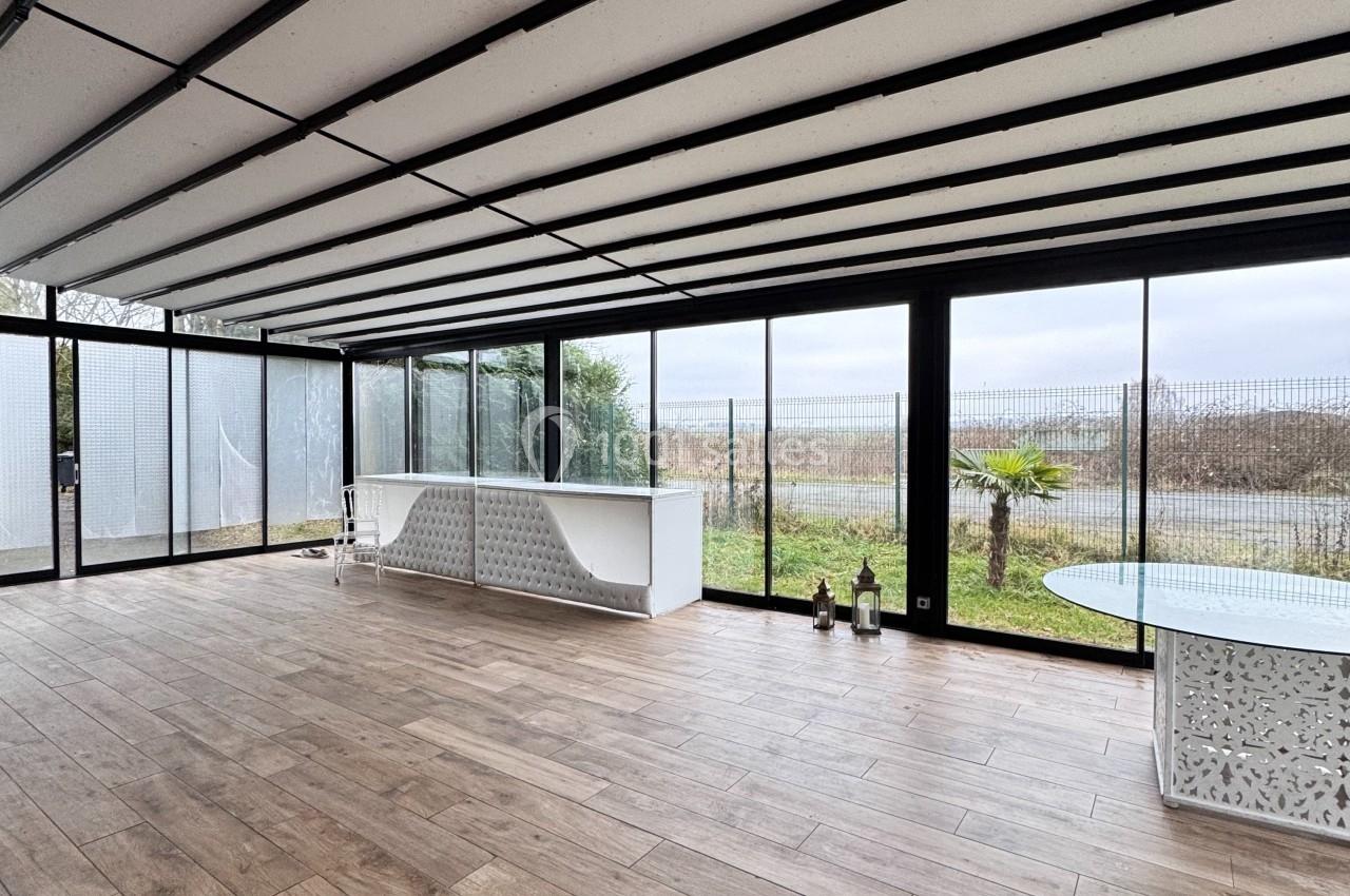 Véranda lumineuse avec sol en bois, grandes baies vitrées donnant sur un jardin et un paysage dégagé.