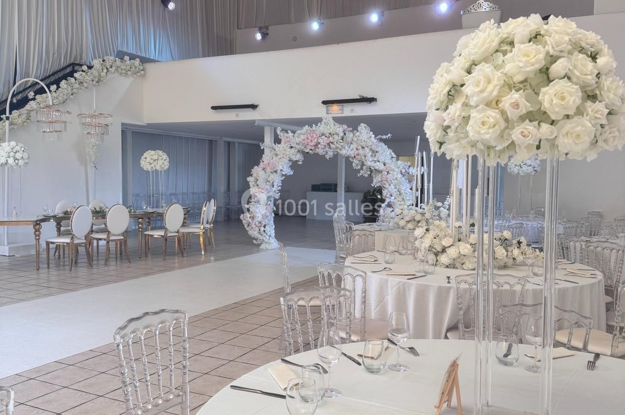 Salle de réception élégante avec tables dressées, chaises transparentes et décorations florales blanches.