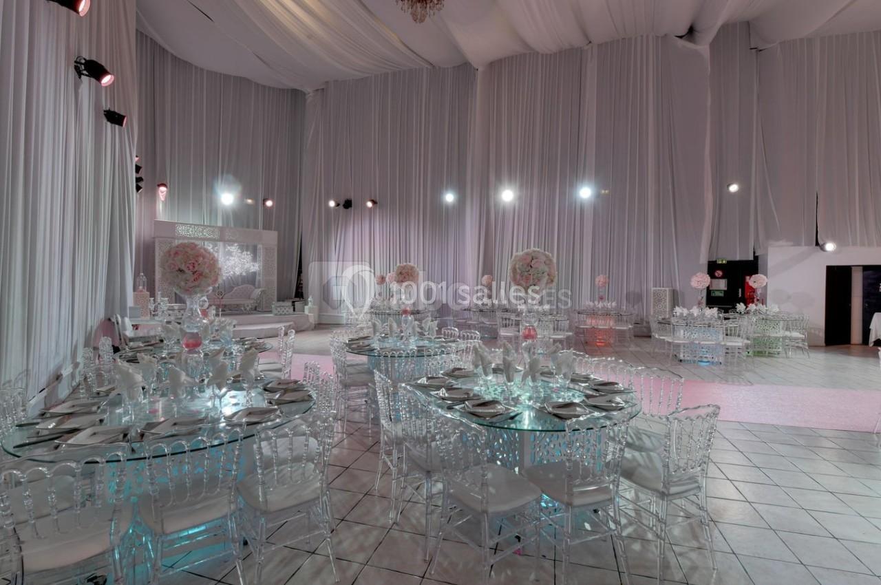 Salle de réception décorée avec des tables rondes dressées, des chaises transparentes et des arrangements floraux roses.