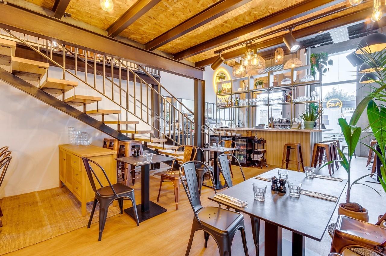 Salle de restaurant lumineuse avec tables en bois, chaises métalliques, bar en fond et escalier en bois sur la gauche.