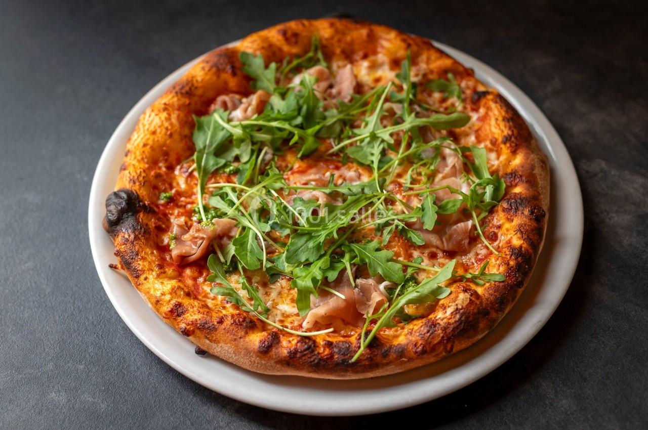 Pizza garnie de roquette, jambon cru et sauce tomate, servie sur une assiette blanche.