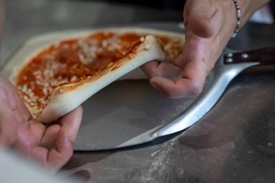 Une pâte à pizza soulevée pour montrer son élasticité, avec une garniture de sauce tomate et fromage visible.