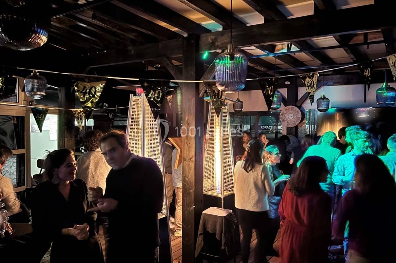 Des personnes discutent et dansent dans un espace intérieur éclairé par des lampes chauffantes et des lumières colorées.