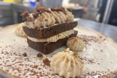 Entrée d'un café avec une grande terrasse, un parasol orange et des meubles en osier sous un ciel bleu.