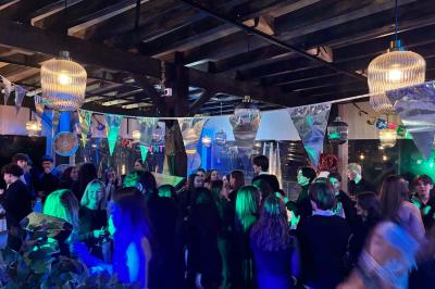 Entrée d'un café avec une grande terrasse, un parasol orange et des meubles en osier sous un ciel bleu.