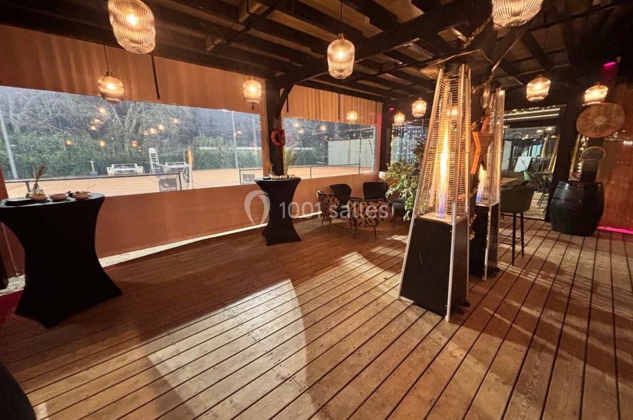 Espace extérieur couvert avec parquet en bois, tables hautes, lampes suspendues et chauffages, vue sur un terrain de sport.