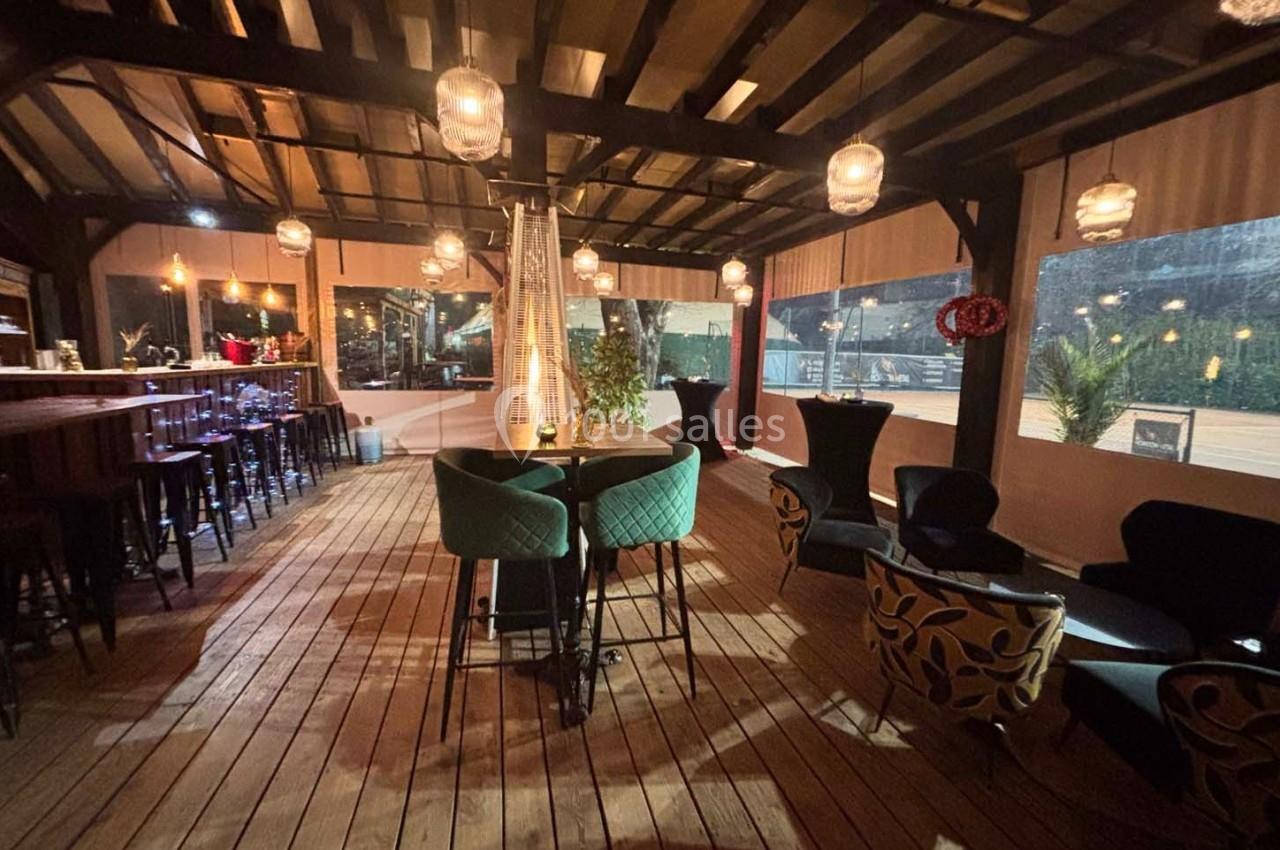 Terrasse couverte en bois avec tables, chaises et éclairage suspendu, ambiance chaleureuse en soirée.