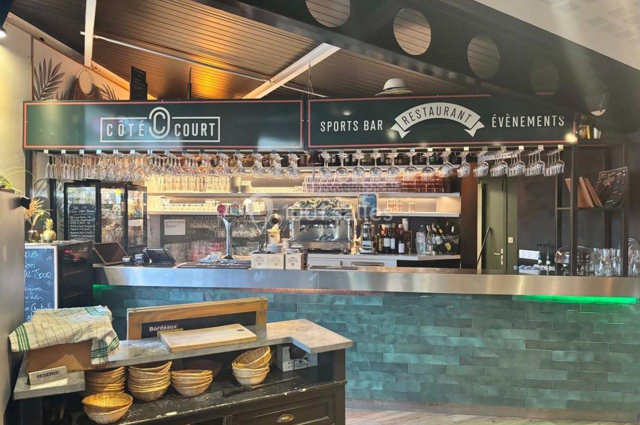 Vue d'un bar-restaurant avec comptoir, étagères de verres suspendus et enseigne ’Côté Court Sports Bar Restaurant Événements…