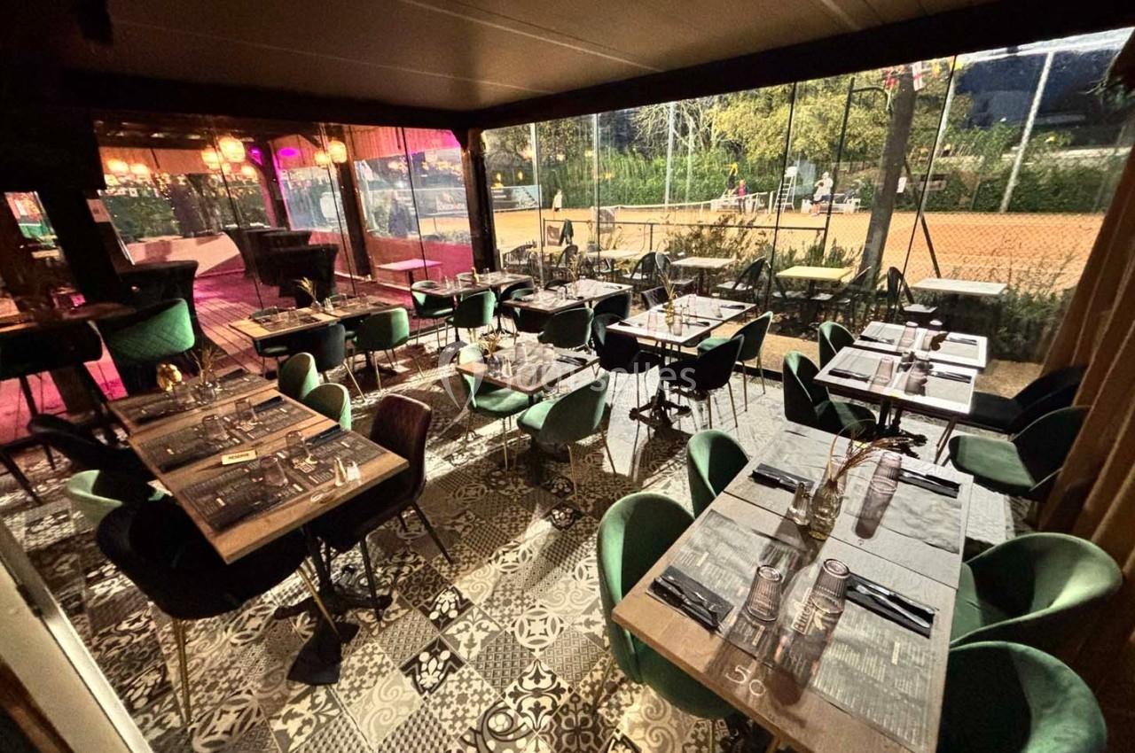 Salle de restaurant élégante avec tables dressées, chaises vertes et vue sur un espace extérieur éclairé.