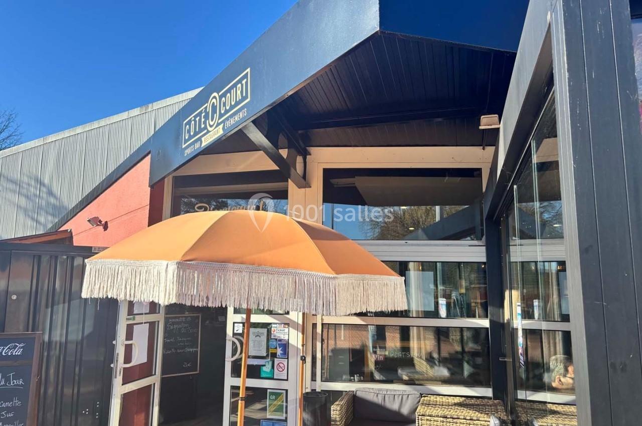 Entrée d'un café avec une terrasse équipée d'un parasol orange et des fauteuils en osier, sous un ciel ensoleillé.