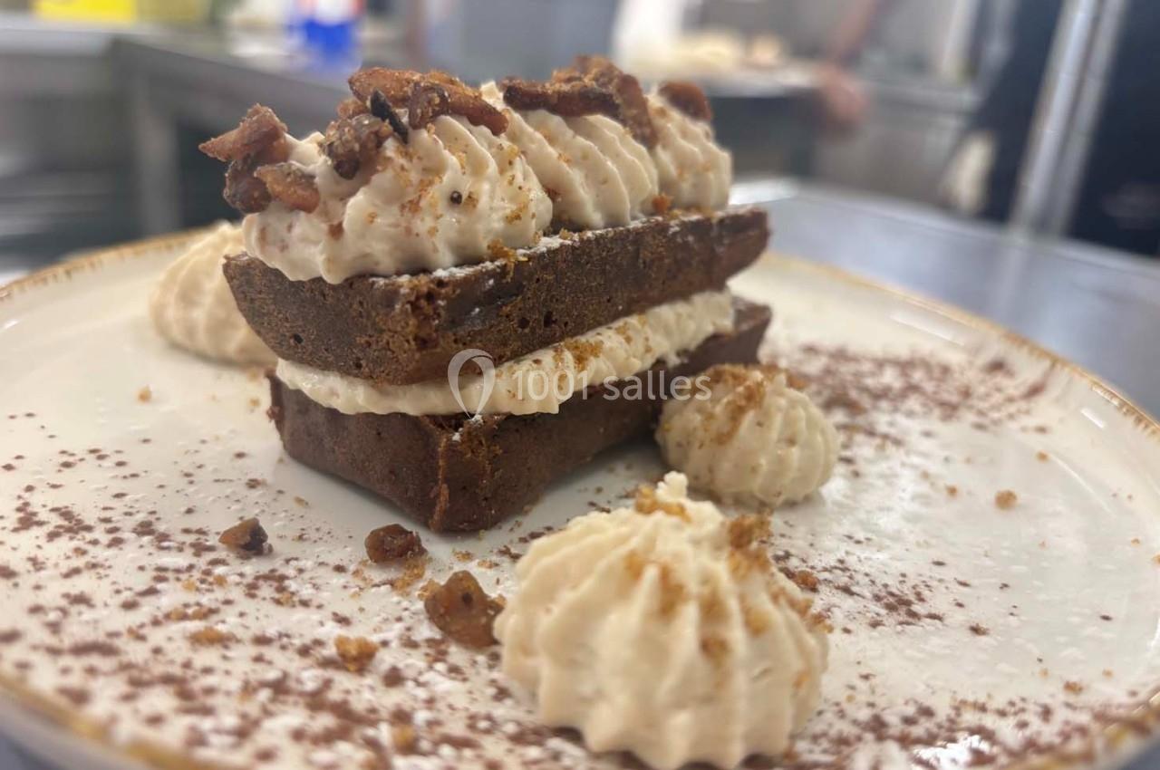 Gâteau au chocolat garni de crème fouettée et éclats croustillants, présenté sur une assiette saupoudrée de cacao.