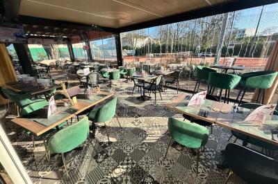 Entrée d'un café avec une terrasse équipée d'un parasol orange et des fauteuils en osier, sous un ciel ensoleillé.