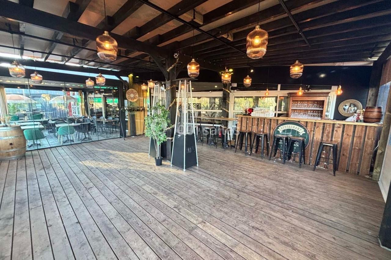 Terrasse en bois couverte avec bar, tabourets hauts, lampes suspendues et tables extérieures visibles en arrière-plan.