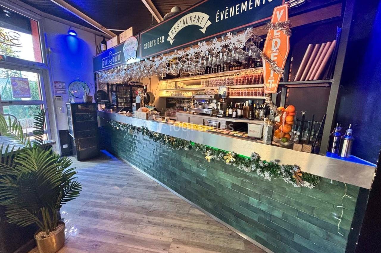 Comptoir de bar décoré avec des guirlandes, étagères remplies de bouteilles et ambiance lumineuse chaleureuse.