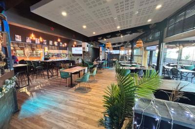 Entrée d'un café avec une terrasse équipée d'un parasol orange et des fauteuils en osier, sous un ciel ensoleillé.