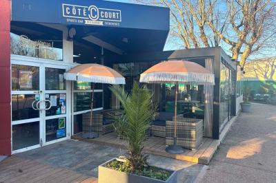 Entrée d'un café avec une terrasse équipée d'un parasol orange et des fauteuils en osier, sous un ciel ensoleillé.