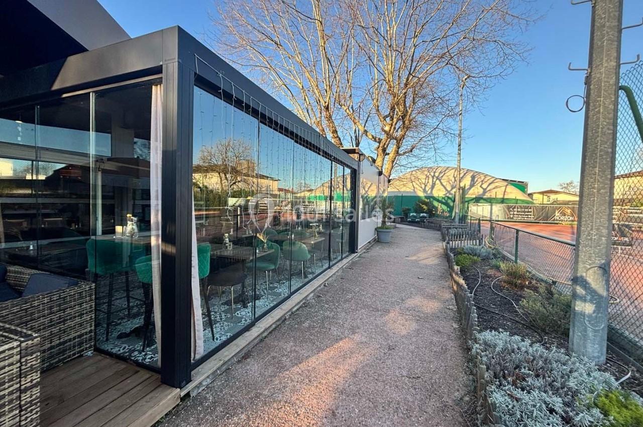 Terrasse vitrée d'un restaurant donnant sur un chemin en gravier, avec vue sur des terrains de tennis et un arbre dénudé.