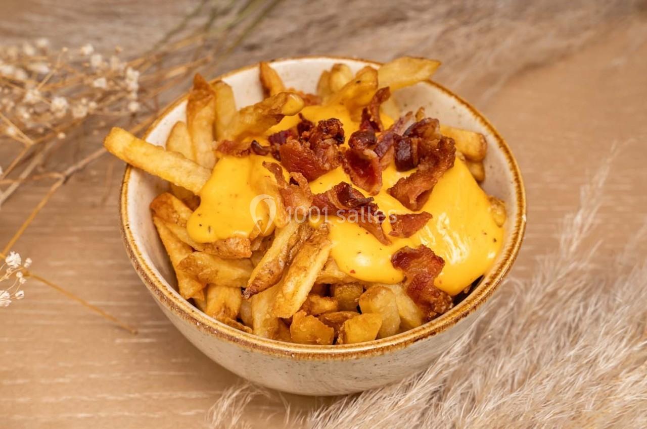 Bol de frites garnies de fromage fondu et de morceaux de bacon, posé sur une table en bois avec des décorations sèches.