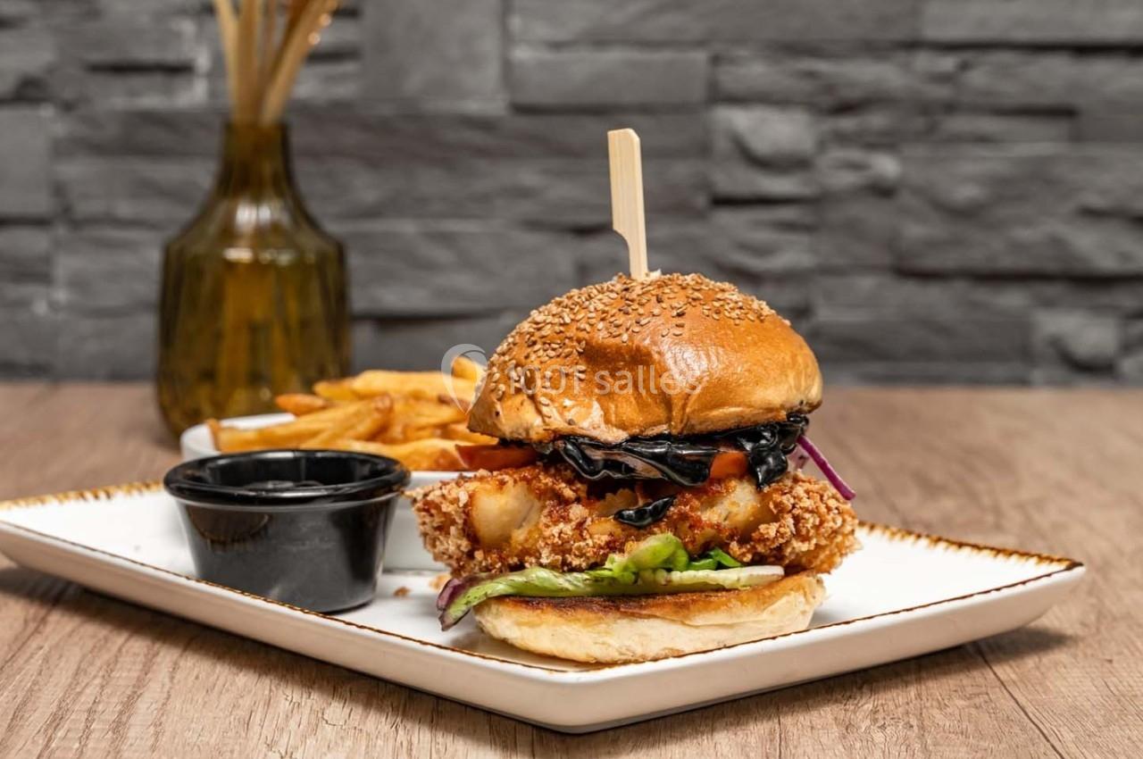 Burger au poulet pané avec légumes, accompagné de frites et d'une sauce, servi sur une assiette blanche.