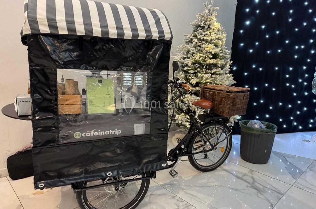 Triporteur noir avec une bâche rayée, stationné devant un sapin décoré et un mur illuminé.