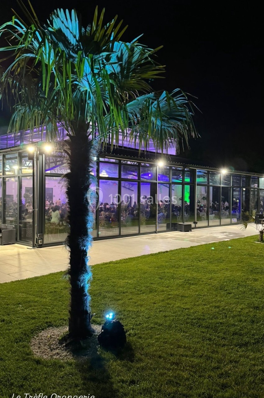 Vue nocturne d'une orangerie vitrée éclairée, entourée de pelouse et d'un palmier au premier plan.