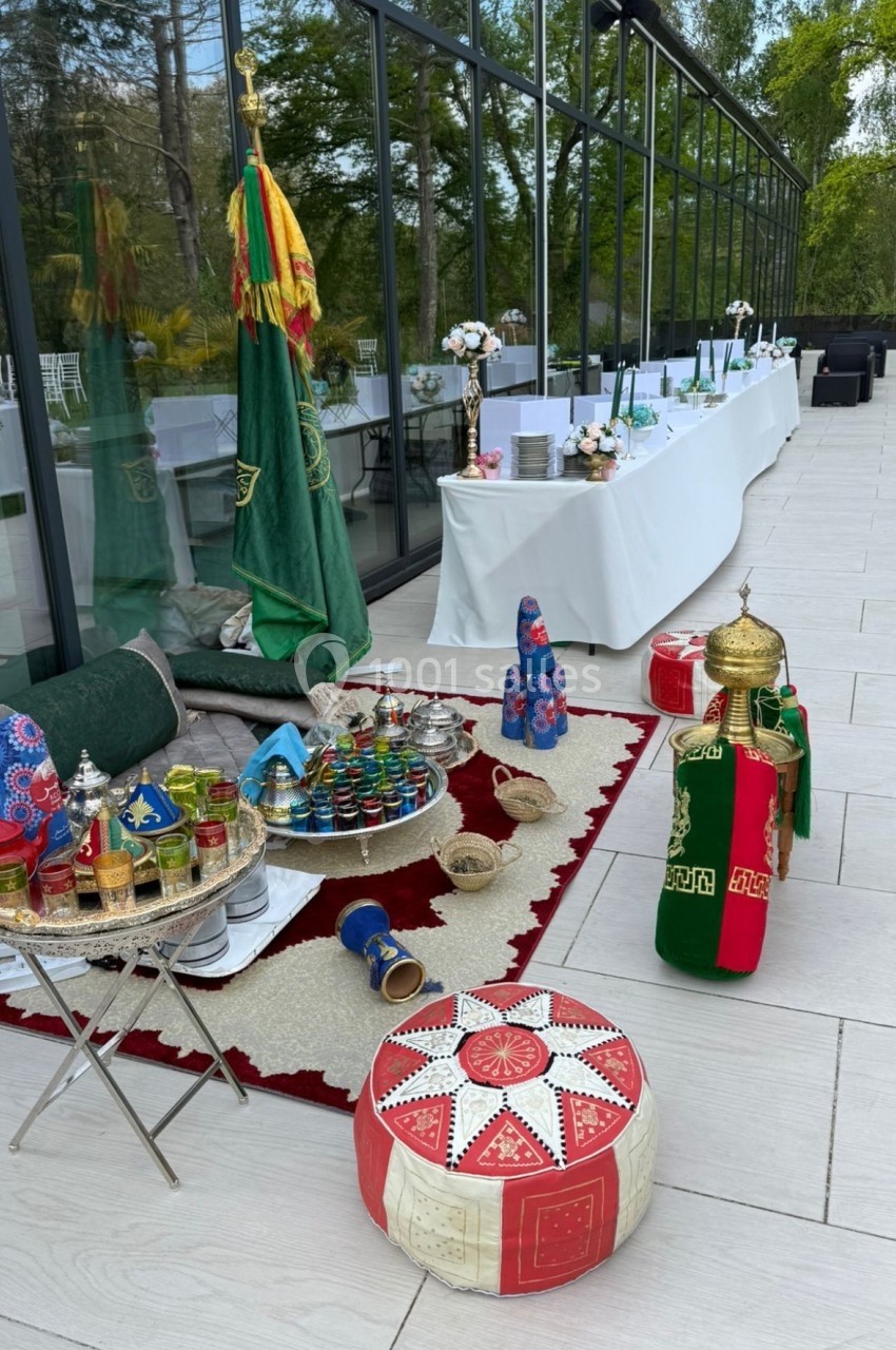 Décoration orientale avec tapis, poufs colorés, lanternes et plateaux de thé, devant une salle vitrée donnant sur un jardin.
