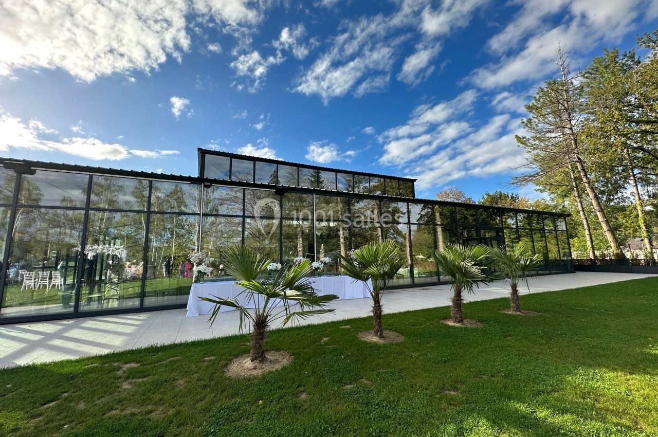 Serre moderne en verre entourée de pelouse, avec des palmiers au premier plan et un ciel partiellement nuageux.