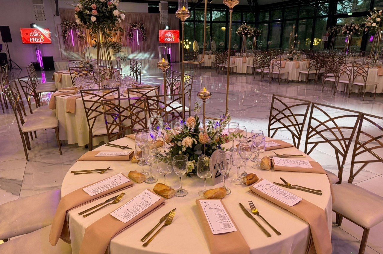 Table de réception élégamment dressée avec nappes beiges, menus, fleurs et couverts dorés dans une salle décorée.