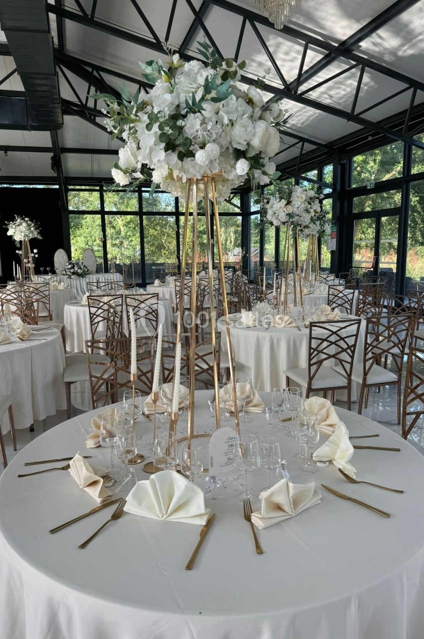 Table de réception élégante avec nappes blanches, serviettes pliées, couverts dorés et centre de table floral sur pied haut.