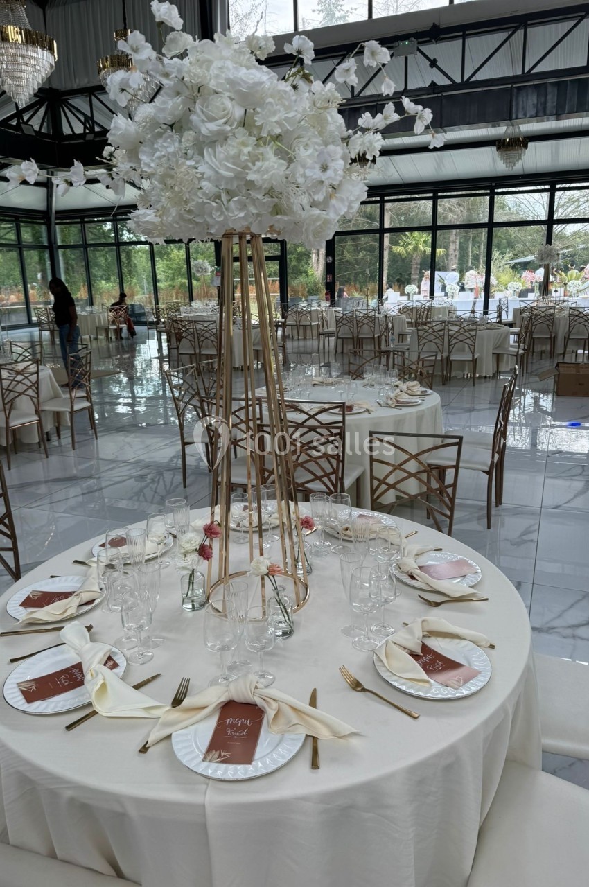Salle de réception élégante avec tables rondes dressées, décorations florales blanches et grandes baies vitrées.