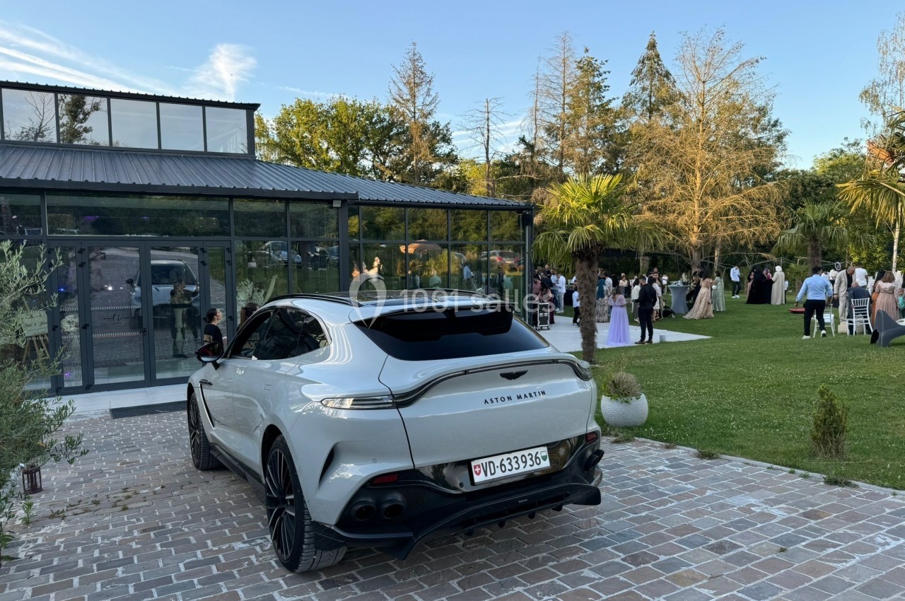 Voiture grise garée devant un bâtiment vitré, avec des invités rassemblés dans un jardin à l'arrière-plan.